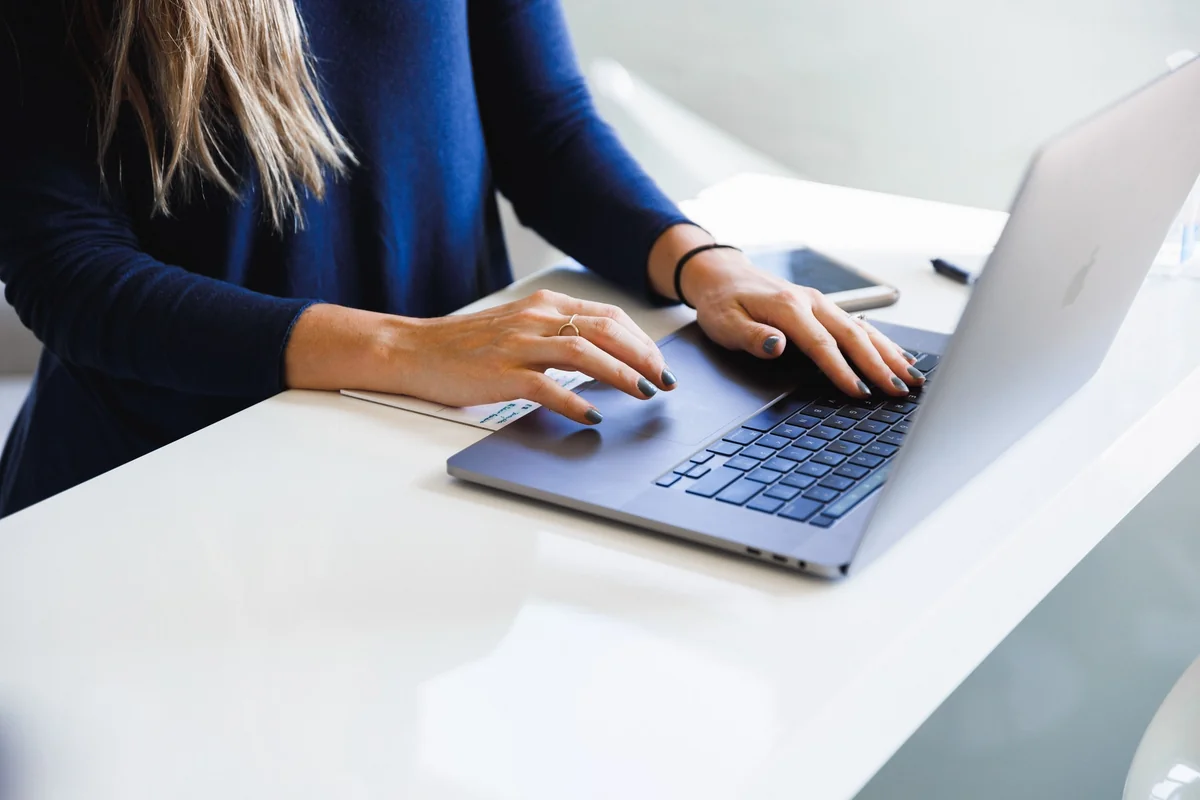 Woman in blue typing on a laptop at a white desk, with a smartphone and pen nearby. Bright setting, focused mood.