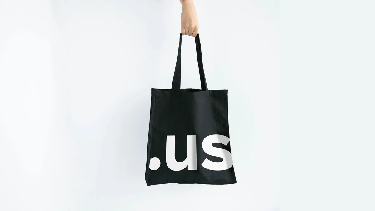 Mano sosteniendo una bolsa negra con texto ".us" en blanco sobre un fondo liso y claro. Diseño limpio y minimalista.